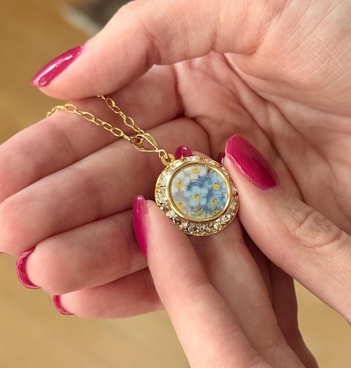 May include: A gold-colored necklace with a round pendant featuring a floral design of blue and yellow flowers. The pendant is encircled by a row of clear crystals. The necklace is held in a person's hands.
