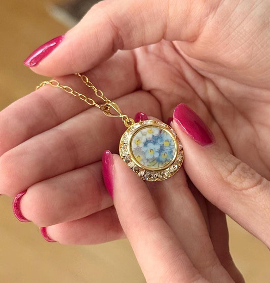 May include: A gold-colored necklace with a round pendant featuring a floral design of blue and yellow flowers. The pendant is encircled by a row of clear crystals. The necklace is held in a person's hands.