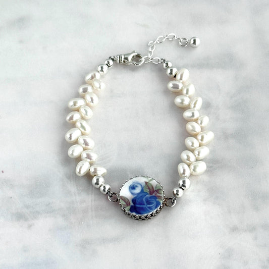May include: A delicate bracelet featuring white freshwater pearls and a central porcelain charm. The charm showcases a blue rose design with green leaves, set in a silver-toned metal frame. The bracelet has a silver clasp and adjustable chain.