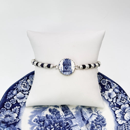 May include: A bracelet featuring a round charm with a blue and white clock tower design. The bracelet has black and silver beads. The bracelet is displayed on a white pillow, with a blue and white floral plate in the background.