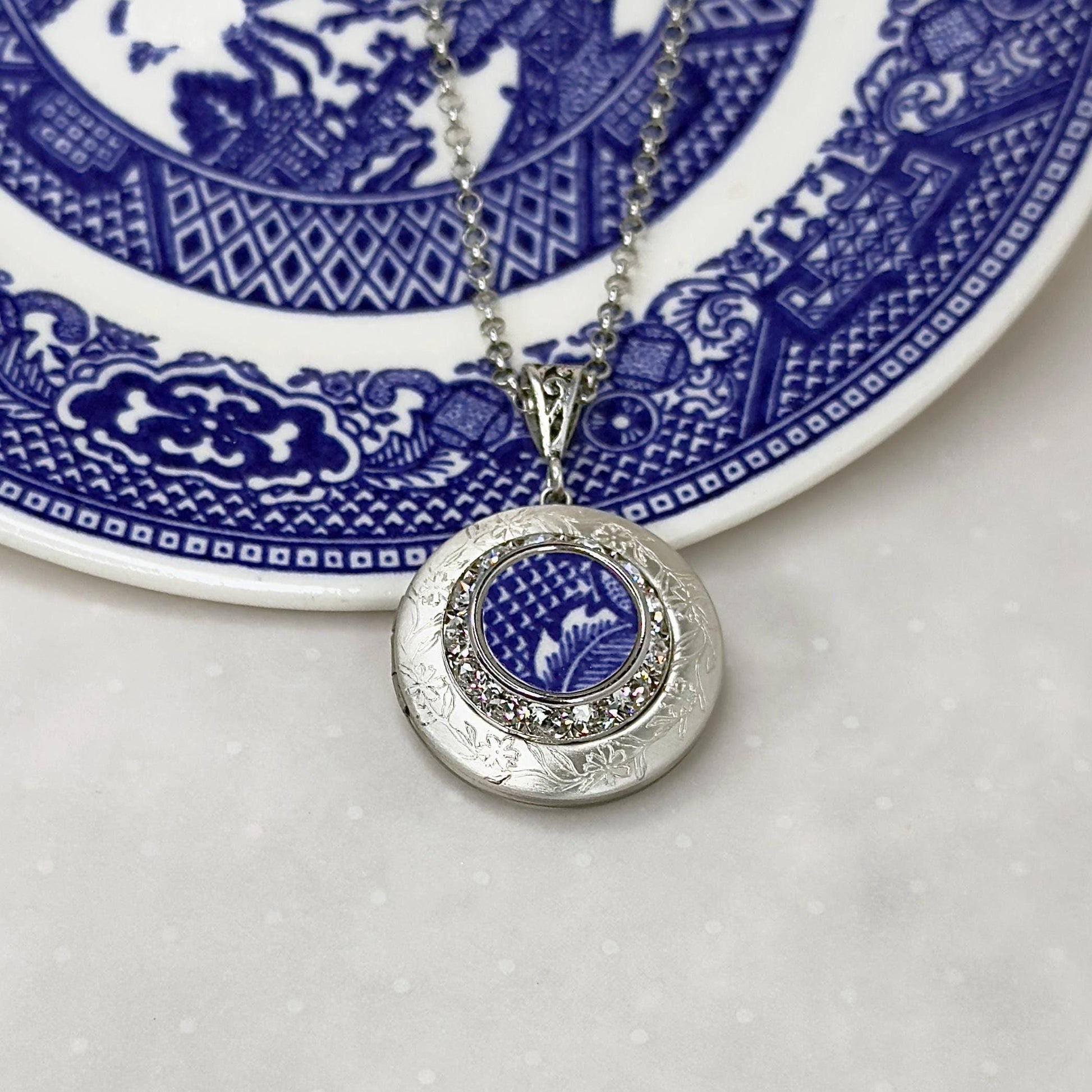May include: A silver locket necklace with a blue and white patterned design inside. The locket is surrounded by a ring of clear rhinestones.