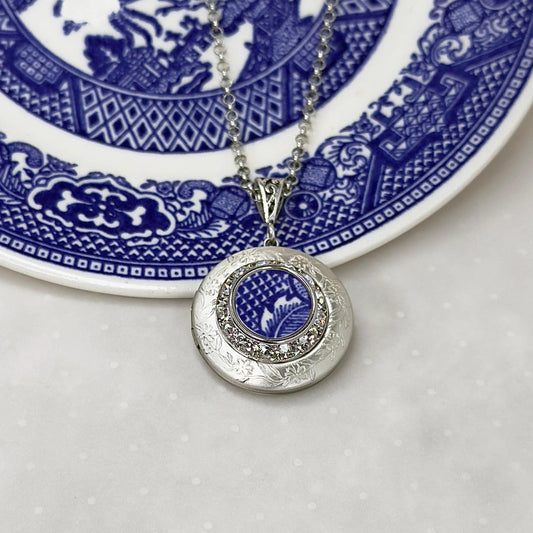 May include: A silver locket necklace with a blue and white patterned design inside. The locket is surrounded by a ring of clear rhinestones.