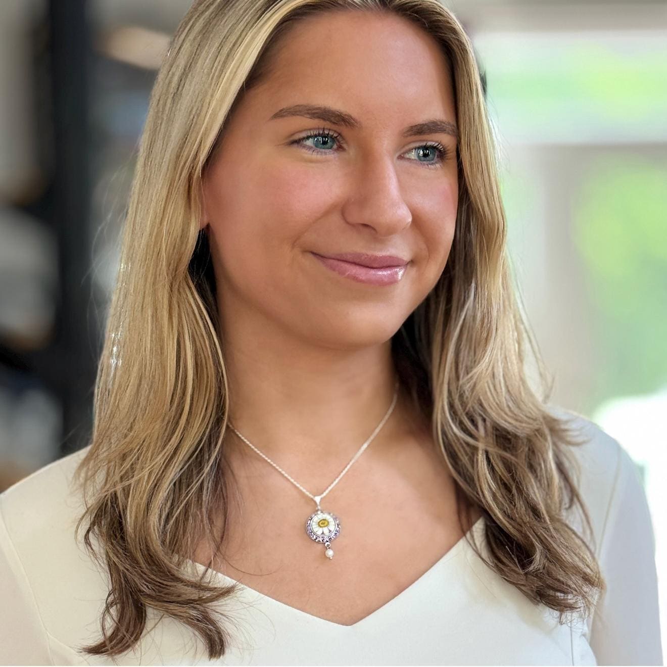 May include: A silver necklace with a daisy pendant. The pendant features a yellow center, white petals, and a small pearl drop. The necklace is worn by a person with blonde hair and blue eyes.