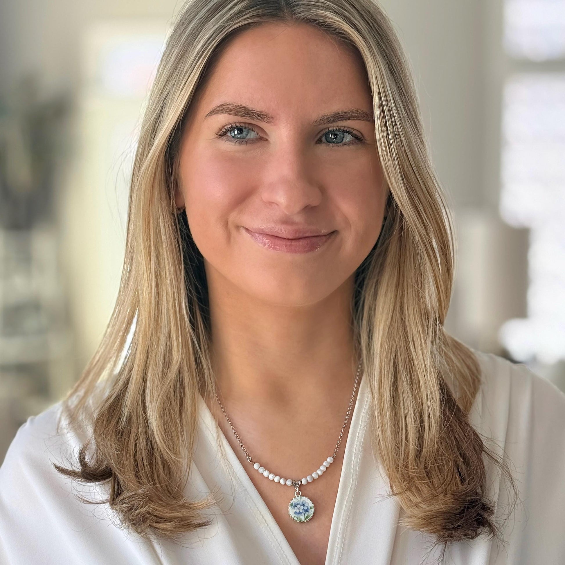 May include: A silver necklace with a floral pendant. The pendant features a detailed blue and white floral design. The necklace has a delicate chain with small, white beads. The woman is wearing a white top.