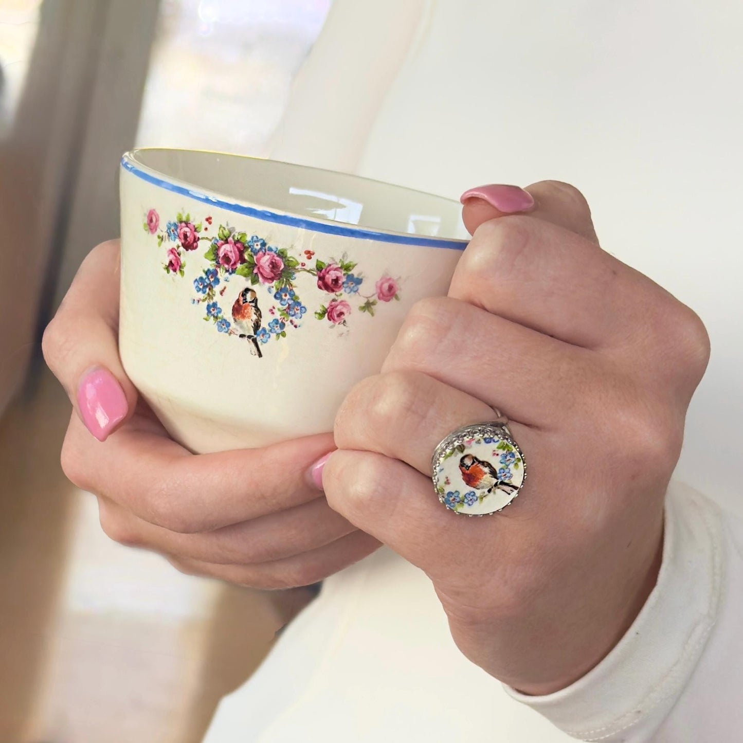 A vintage white ceramic teacup with a blue rim and floral and bird design, held by a person wearing a ring featuring the  bird and floral forget me not design. The teacup has a design of pink roses and blue forget me notflowers