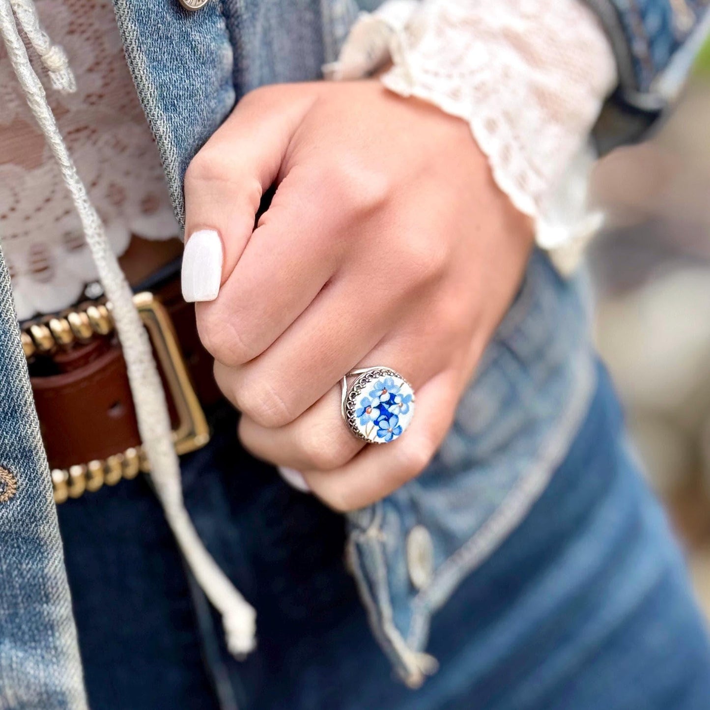 A sterling silver ring with a large, round face featuring a blue forget-me-not floral design. The ring's setting has an filigree border. The ring is worn on a finger, with a denim jacket and white lace sleeve in the background.