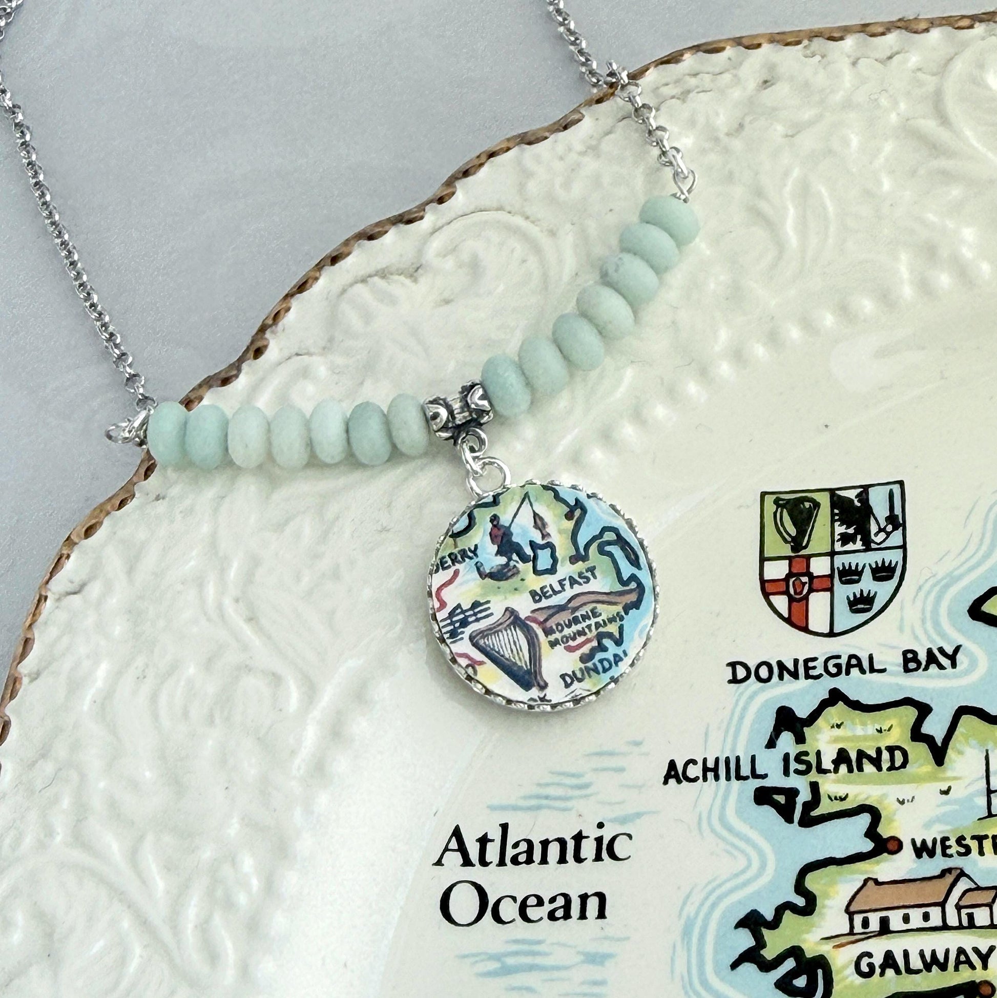 May include: A silver necklace with a pendant featuring a map of Ireland. The pendant is round with a colorful illustration of the country's landmarks. The necklace has light blue beads and is displayed on a decorative plate.