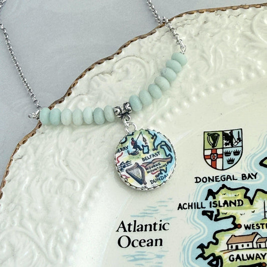 May include: A silver necklace with a pendant featuring a map of Ireland. The pendant is round with a colorful illustration of the country's landmarks. The necklace has light blue beads and is displayed on a decorative plate.