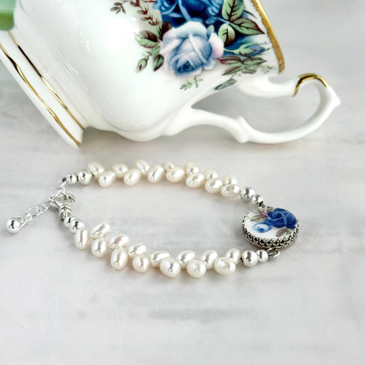 May include: A delicate bracelet featuring white freshwater pearls and a decorative porcelain charm with a blue rose design. The bracelet has a silver-tone chain and clasp. A white teacup with blue floral accents is in the background.