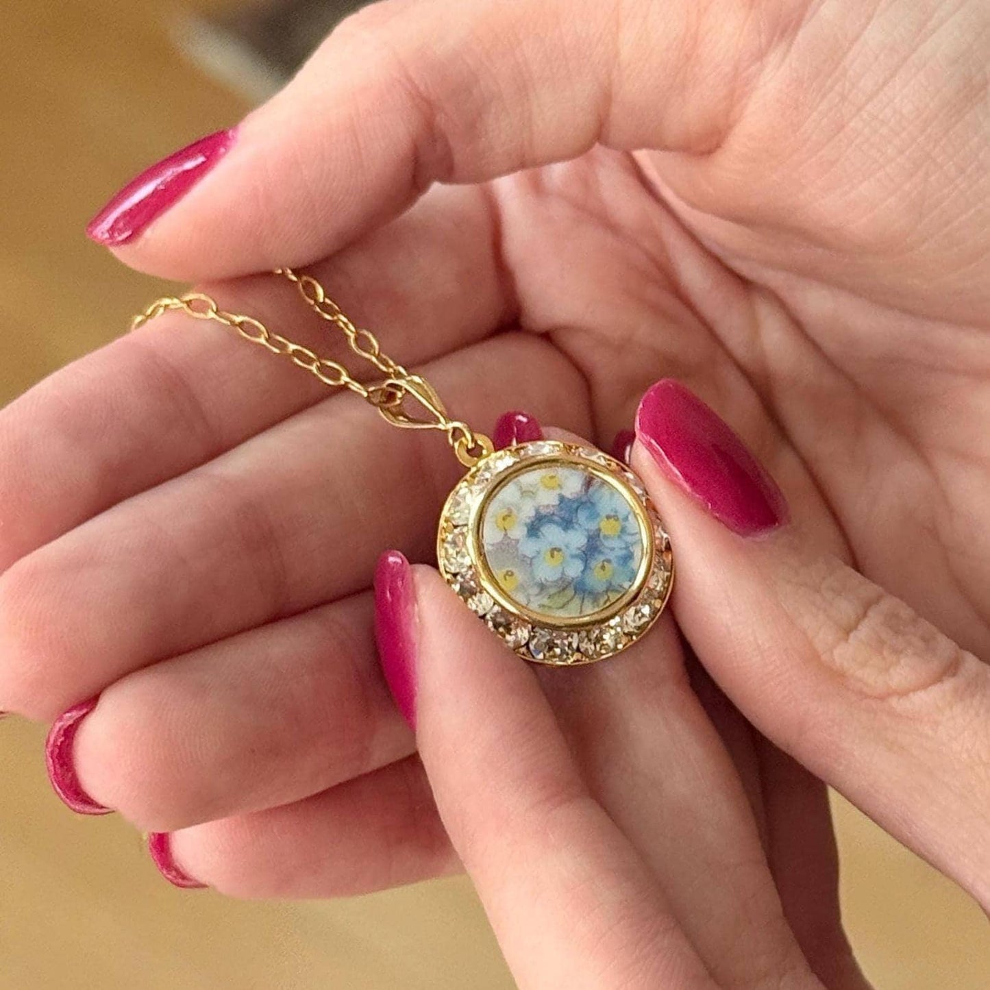 May include: A gold-colored necklace with a round pendant featuring a floral design of blue and yellow flowers. The pendant is encircled by a row of clear crystals. The necklace is held in a person's hands.