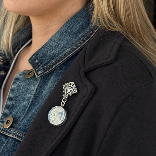 May include: A silver-toned brooch with an ornate top design and a dangling circular pendant. The pendant features a white cameo design with blue accents. The brooch is attached to a black blazer and a denim jacket.