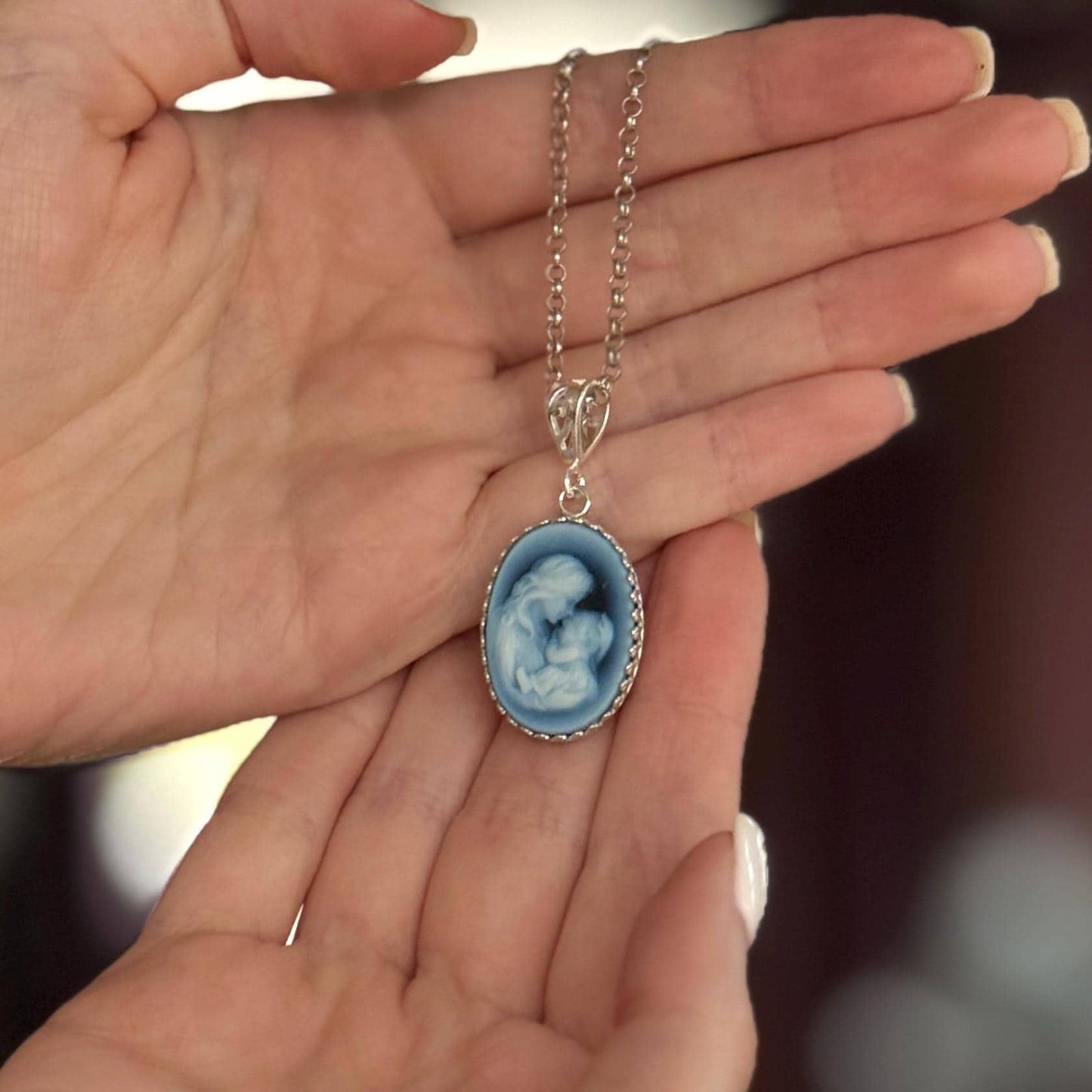 May include: A silver-toned necklace featuring an oval pendant with a blue cameo design. The cameo depicts a mother and child. The pendant is framed with a decorative border and hangs from a delicate chain.