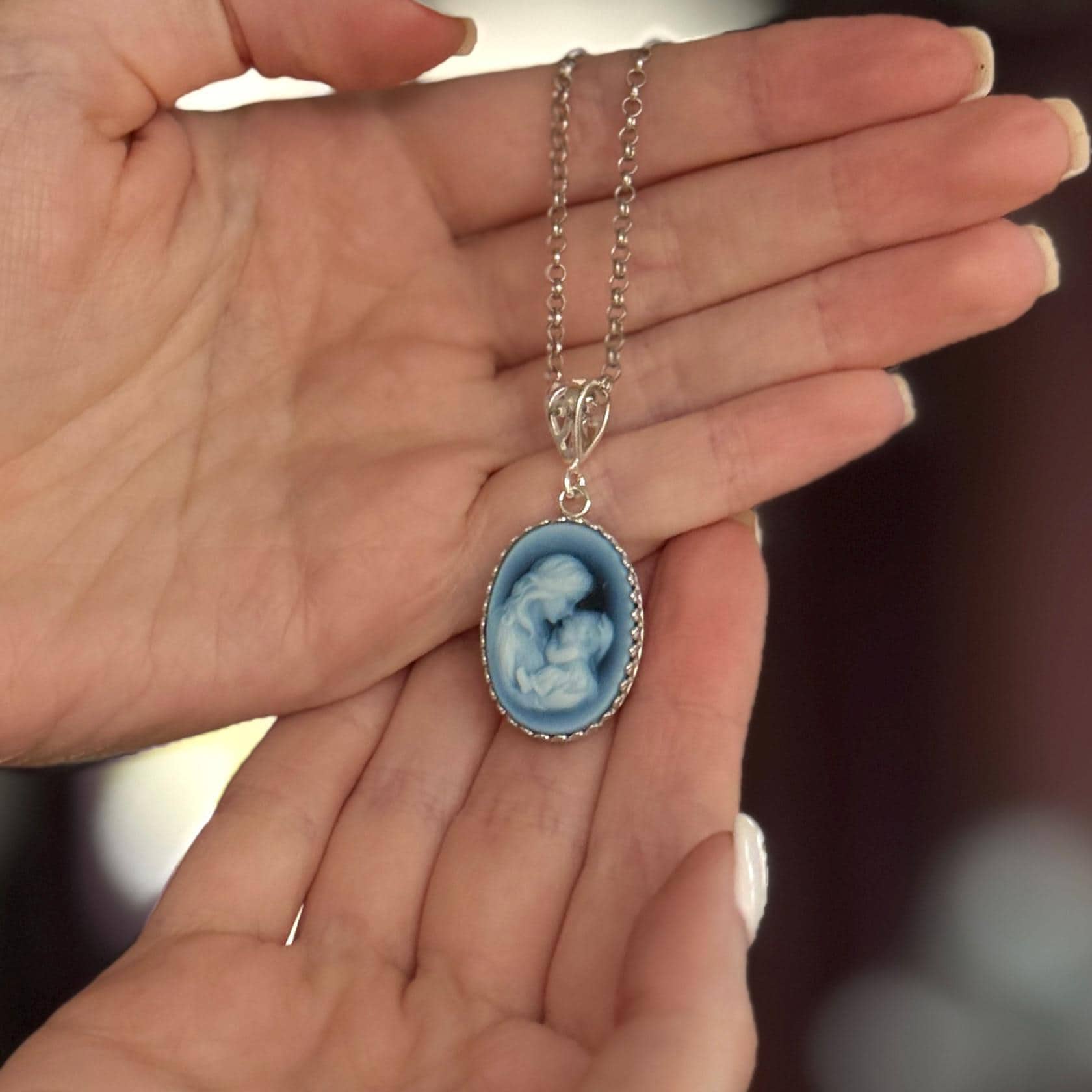 May include: A silver-toned necklace featuring an oval pendant with a blue cameo design. The cameo depicts a mother and child. The pendant is framed with a decorative border and hangs from a delicate chain.