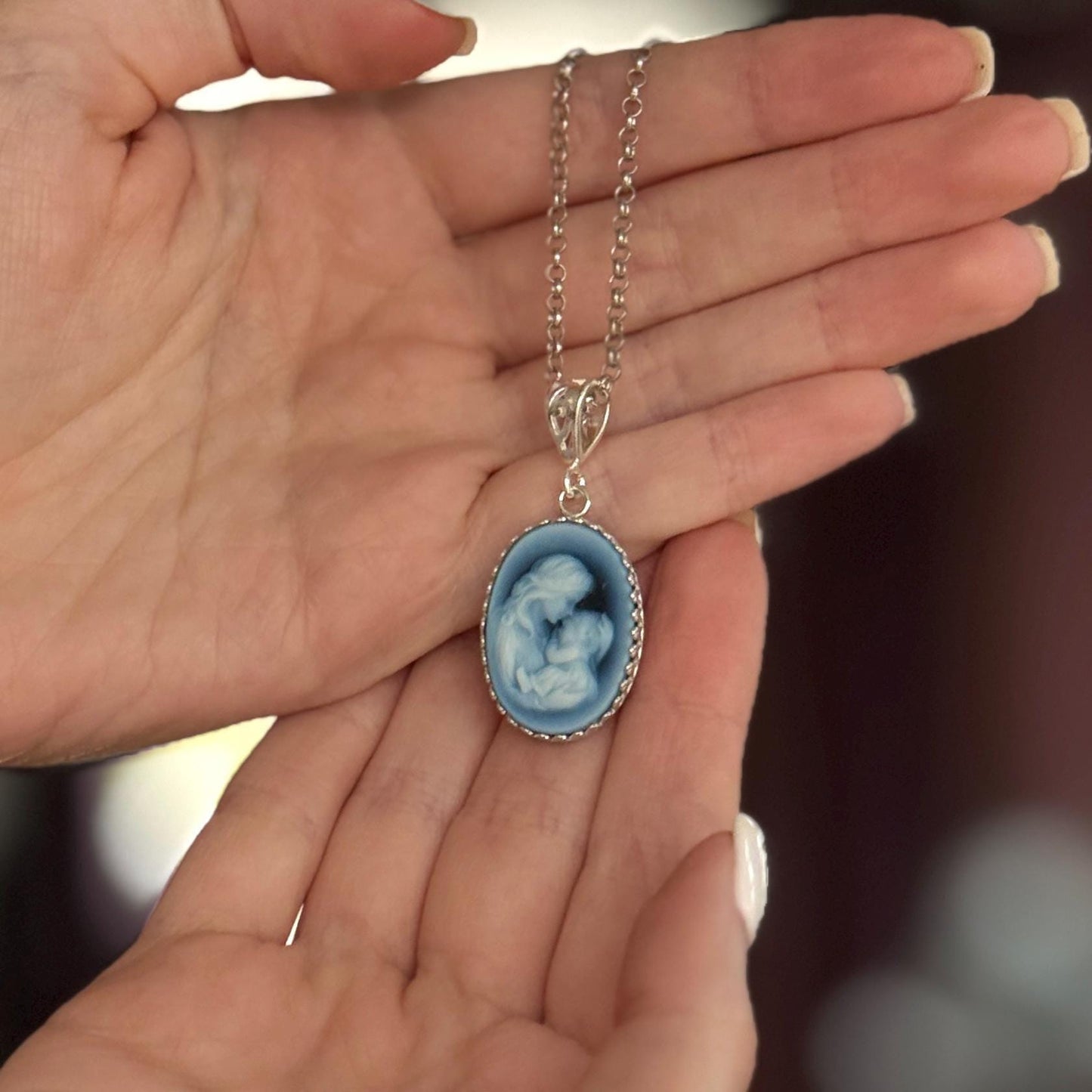 May include: A silver-toned necklace featuring an oval pendant with a blue cameo design. The cameo depicts a mother and child. The pendant is framed with a decorative border and hangs from a delicate chain.