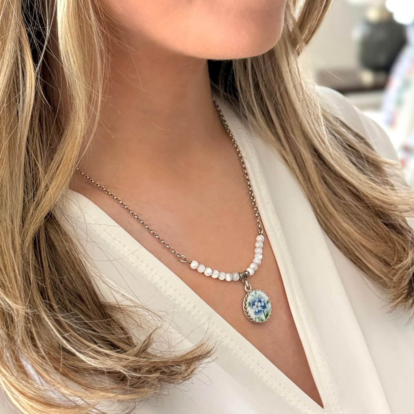 May include: A silver necklace with a pendant featuring a blue floral design. The necklace has a delicate chain and is adorned with small, white beads. The pendant is round with a silver border.