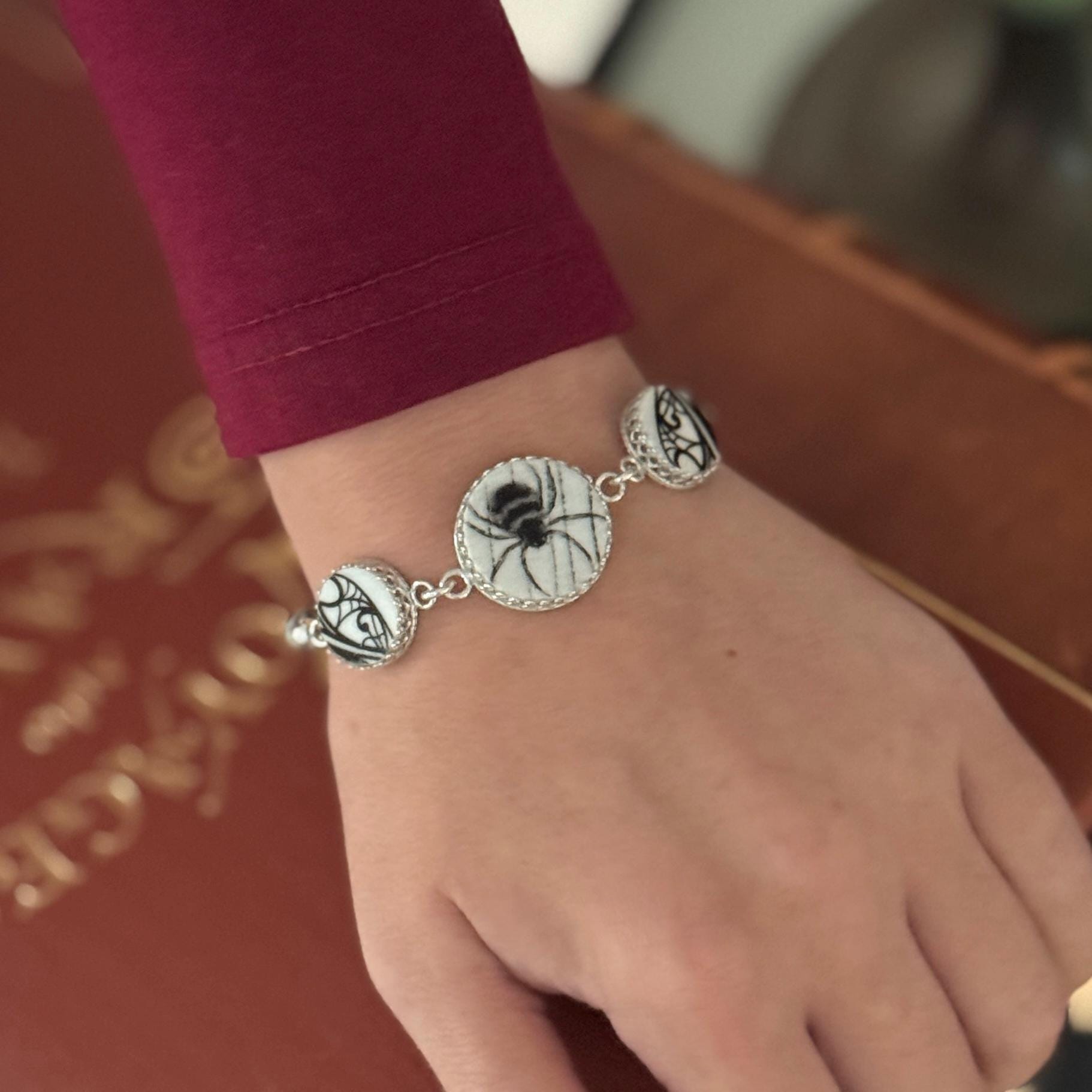 May include: A silver bracelet featuring three circular charms. The central charm displays a black spider on a white background. The other charms have black and white designs. The bracelet is worn on a wrist.