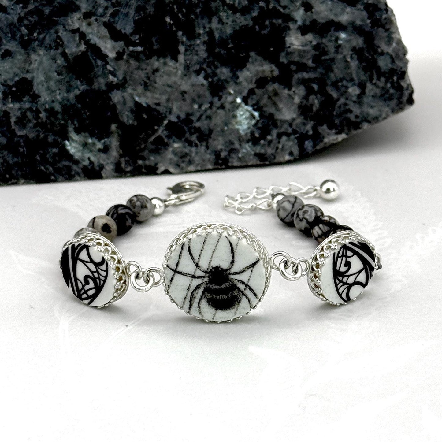 May include: A silver bracelet featuring a central round charm with a black spider design on a white background. The bracelet includes black and gray stone beads and decorative silver accents. The bracelet is designed with a clasp closure.