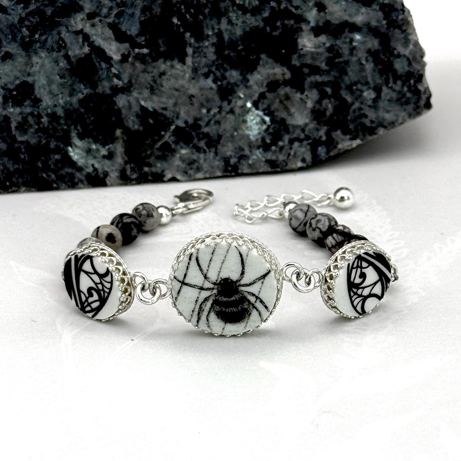 May include: A silver bracelet featuring a central round charm with a black spider design on a white background. The bracelet includes black and gray stone beads and decorative silver accents. The bracelet is designed with a clasp closure.