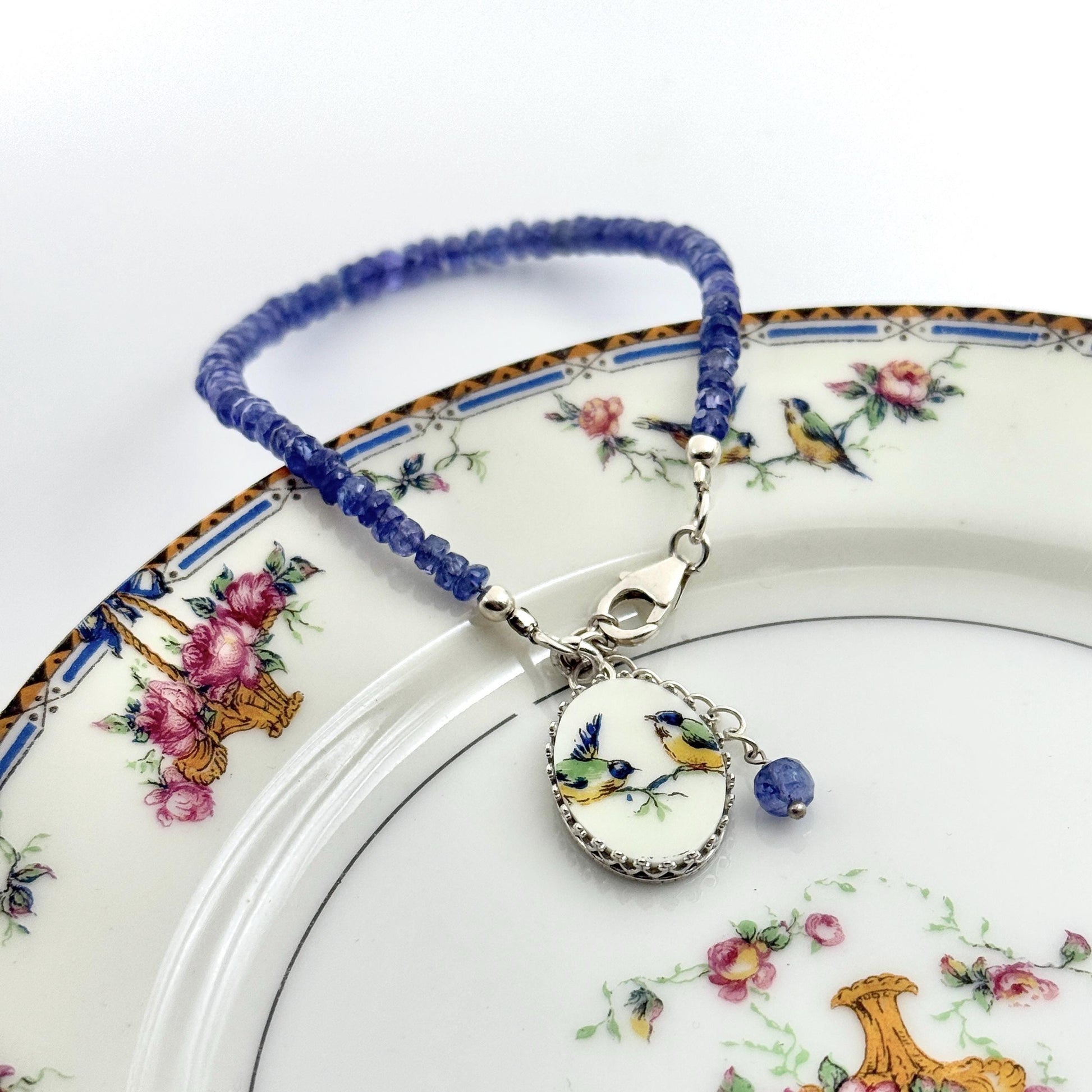 May include: A blue beaded bracelet with a silver clasp and a decorative oval pendant. The pendant features a painted image of two birds on a branch. A small blue bead dangles from the pendant. The bracelet is displayed on a floral patterned plate.