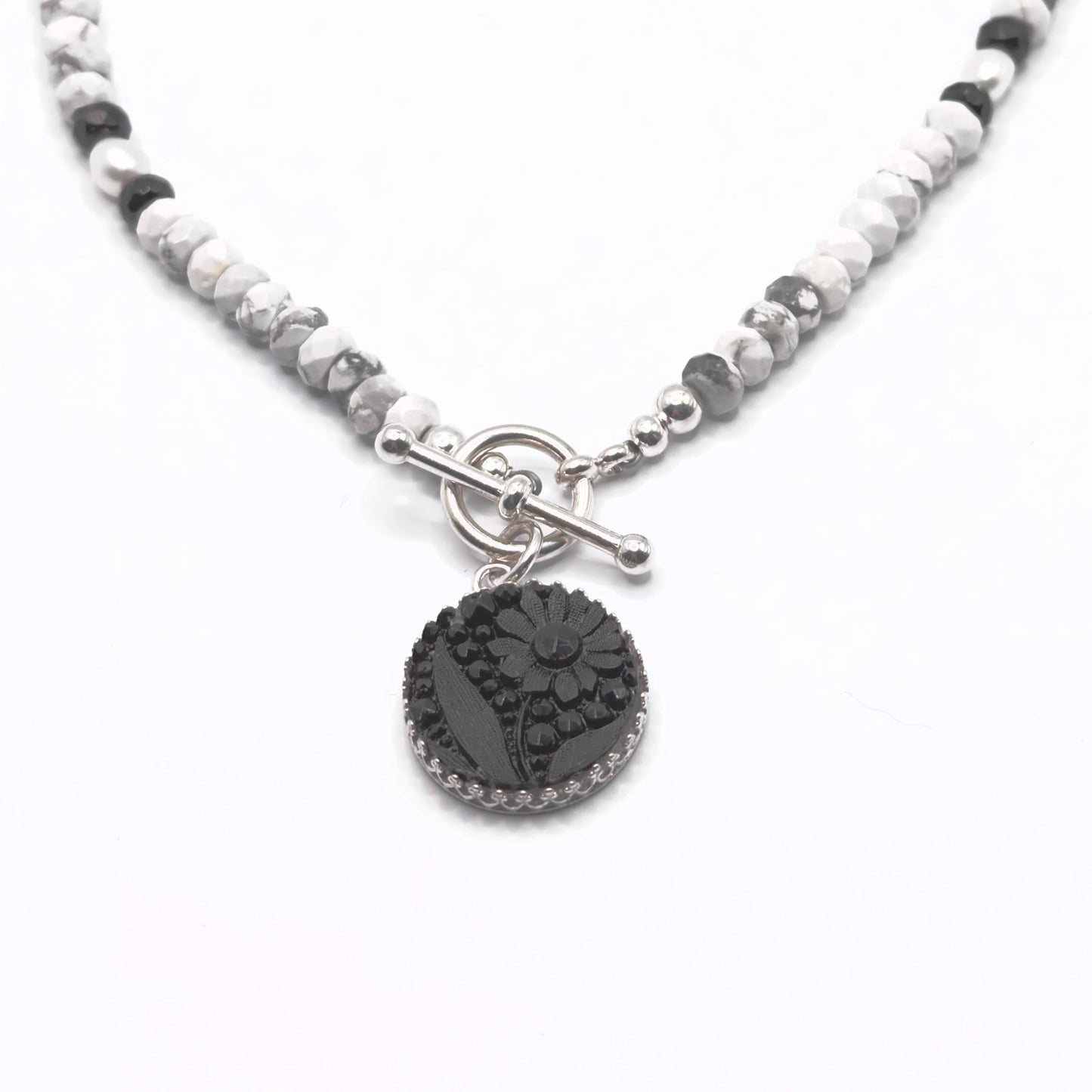 May include: A necklace featuring a black floral pendant with a silver-colored border. The pendant hangs from a beaded chain of white, gray, and black stones. The necklace has a silver-colored toggle clasp.