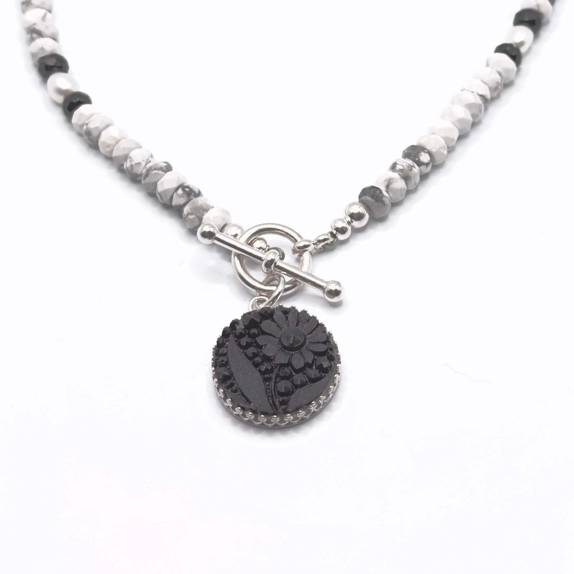 May include: A necklace featuring a black floral pendant with a silver-colored border. The pendant hangs from a beaded chain of white, gray, and black stones. The necklace has a silver-colored toggle clasp.