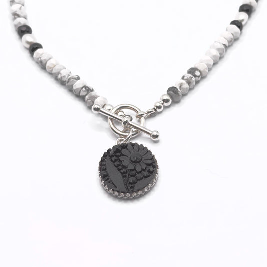 May include: A necklace featuring a black floral pendant with a silver-colored border. The pendant hangs from a beaded chain of white, gray, and black stones. The necklace has a silver-colored toggle clasp.