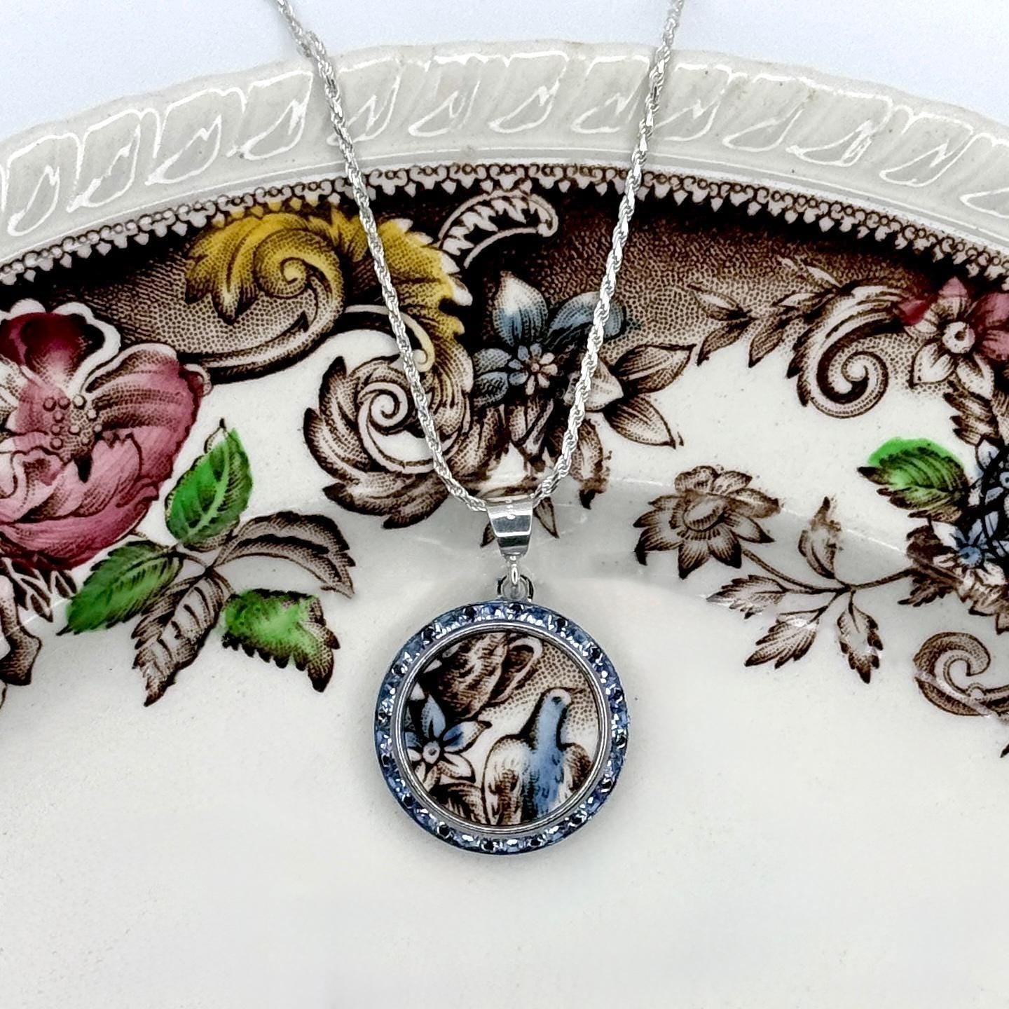 May include: A silver necklace with a round pendant featuring a blue bird design. The pendant is framed with blue crystals and hangs on a delicate chain. The necklace rests on a floral patterned plate.