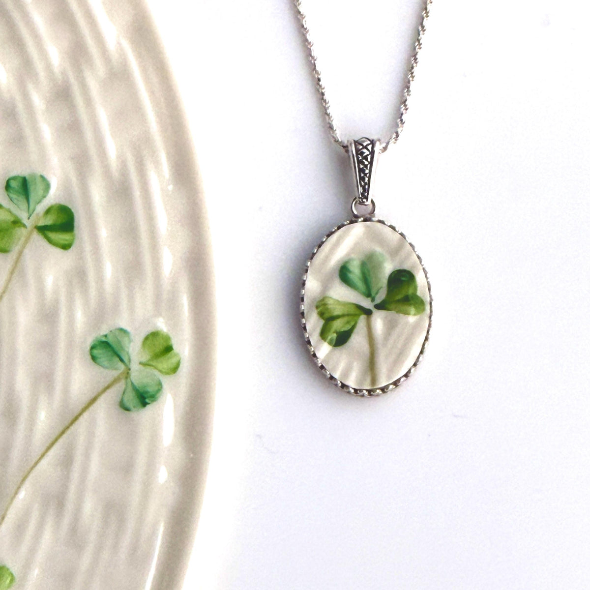 May include: Silver-toned necklace with an oval pendant featuring a hand-painted green shamrock on a cream-colored background. The pendant is framed with a decorative silver border. The necklace is a delicate piece of jewelry.
