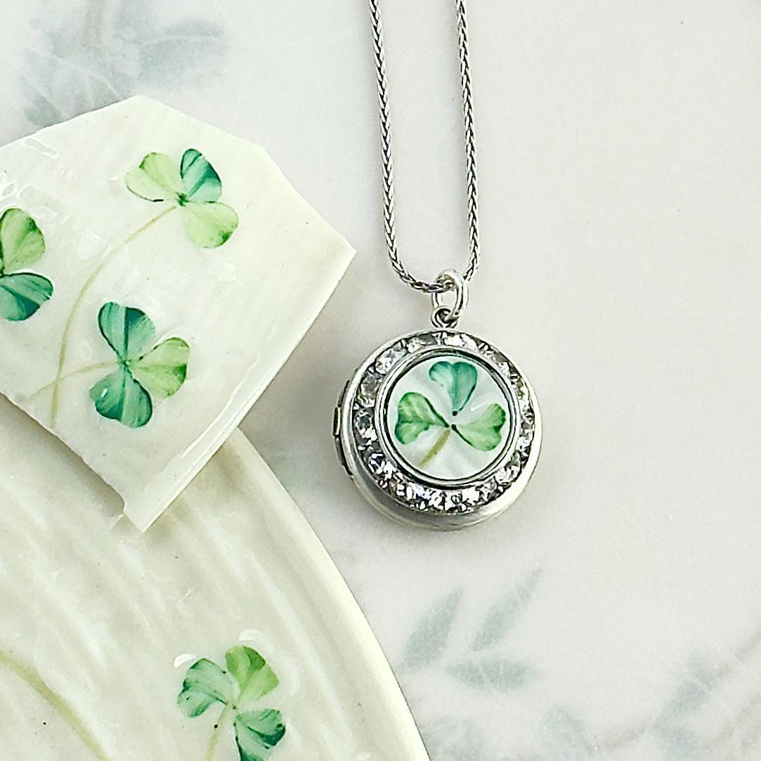 May include: A silver-toned pendant necklace with a round locket featuring a green and white shamrock design. The locket is surrounded by clear crystals. A matching shamrock design is painted on a white ceramic dish.