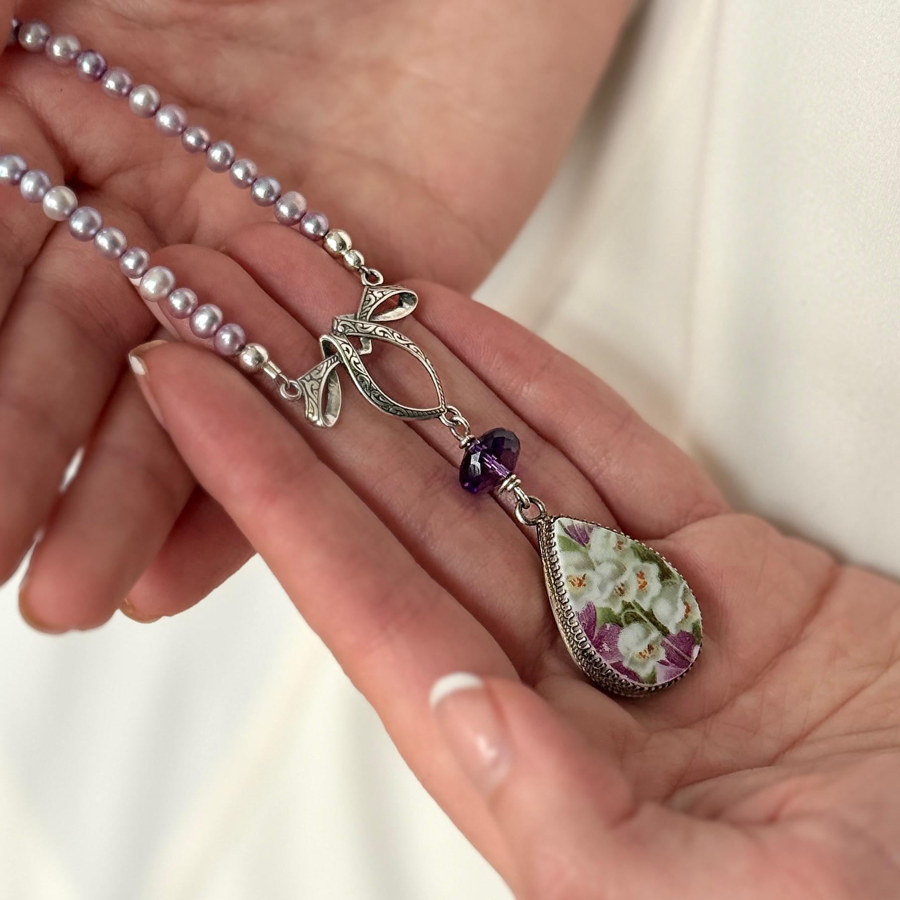 May include: A silver necklace with a teardrop pendant featuring a floral design in white and purple. The necklace has a string of small, light purple pearls and a purple gemstone accent.