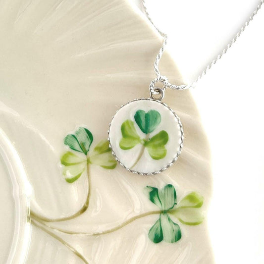 May include: A silver necklace with a round pendant featuring a hand-painted green shamrock design. The pendant has a white background and a decorative silver border. The necklace is displayed on a cream-colored surface with painted shamrock accents.