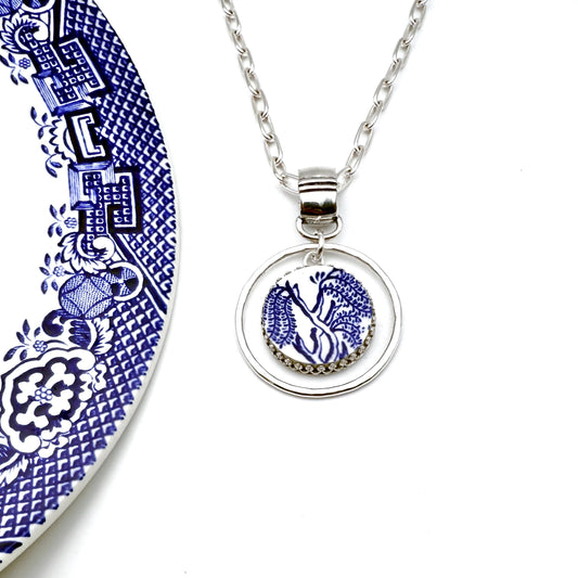 May include: A silver-toned necklace with a circular pendant featuring a blue and white design. The pendant is suspended from a chain and set against a white background. The design on the pendant resembles a floral or botanical pattern.