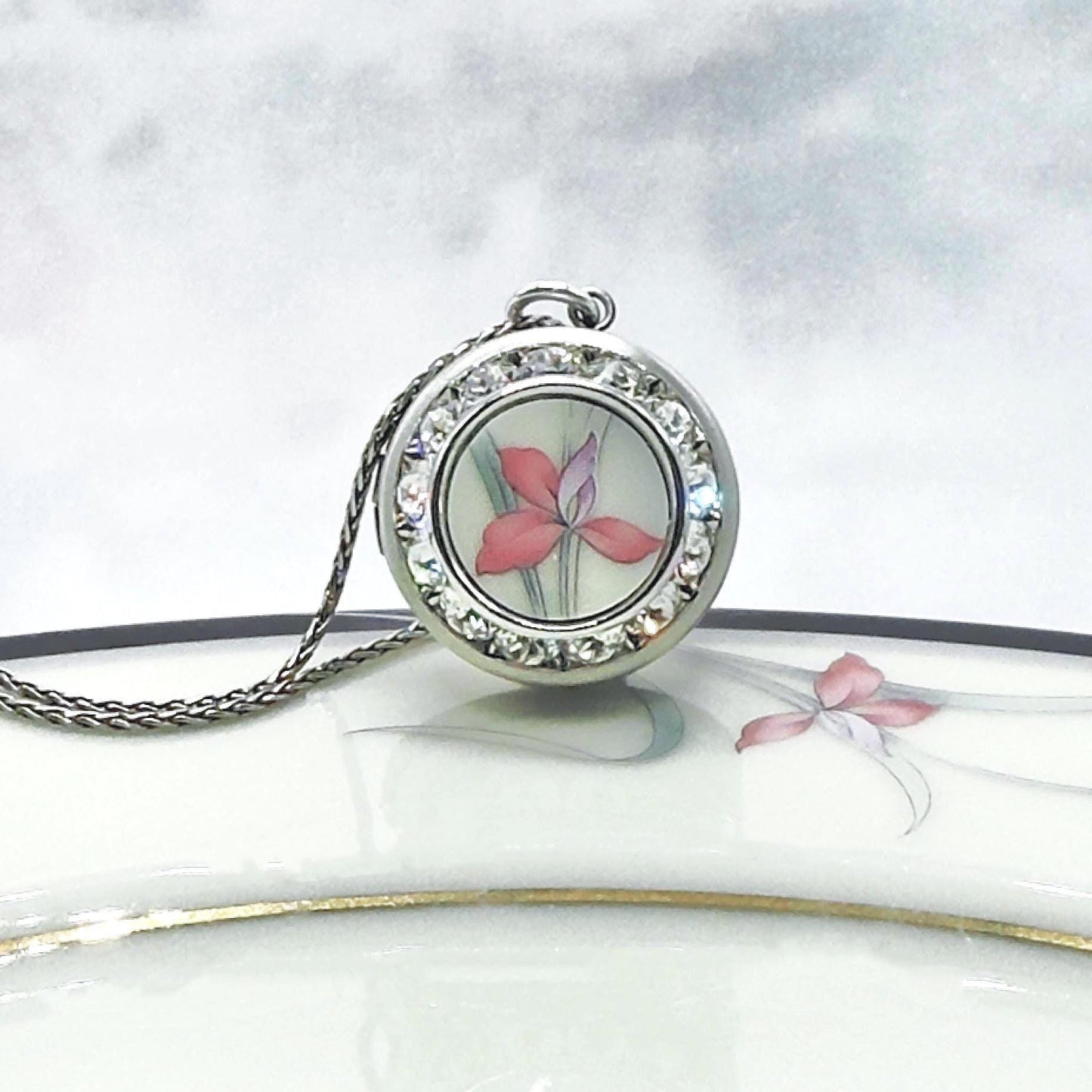 May include: A silver-toned pendant necklace featuring a round locket with a floral design. The locket is adorned with clear crystals around the edge. The floral design inside the locket showcases a pink and purple flower. The necklace chain is also silver-toned.