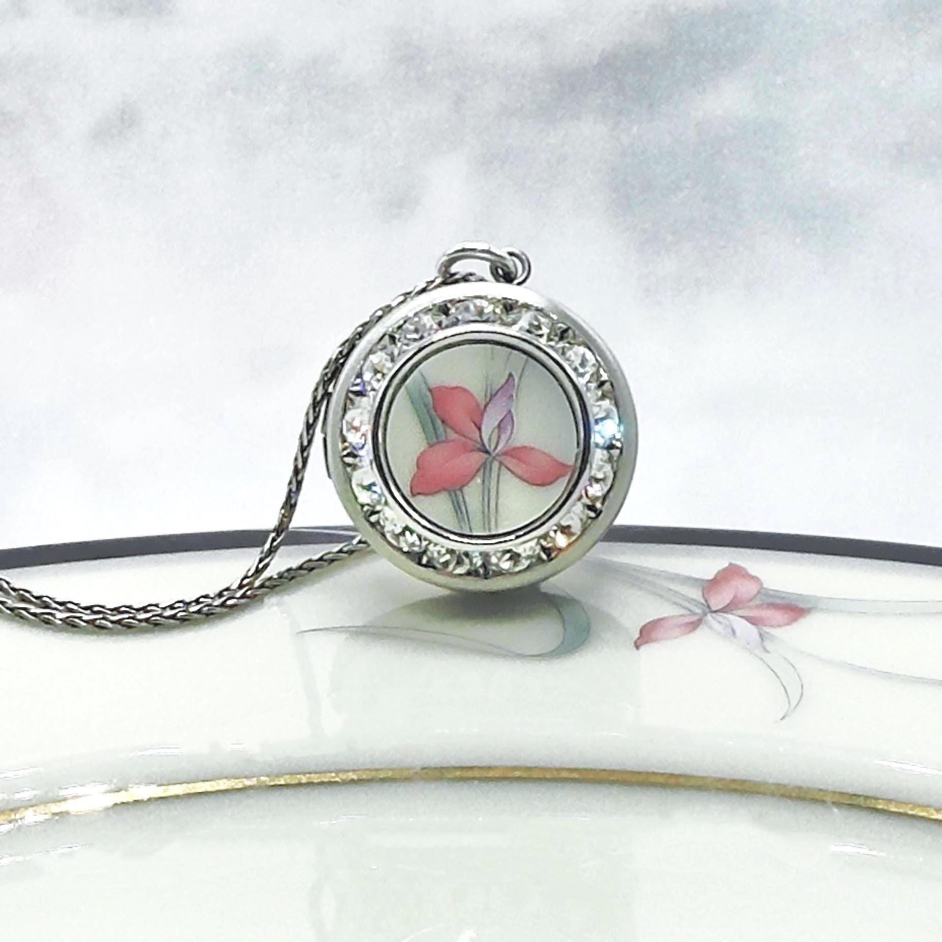 May include: A silver-toned pendant necklace featuring a round locket with a floral design. The locket is adorned with clear crystals around the edge. The floral design inside the locket showcases a pink and purple flower. The necklace chain is also silver-toned.