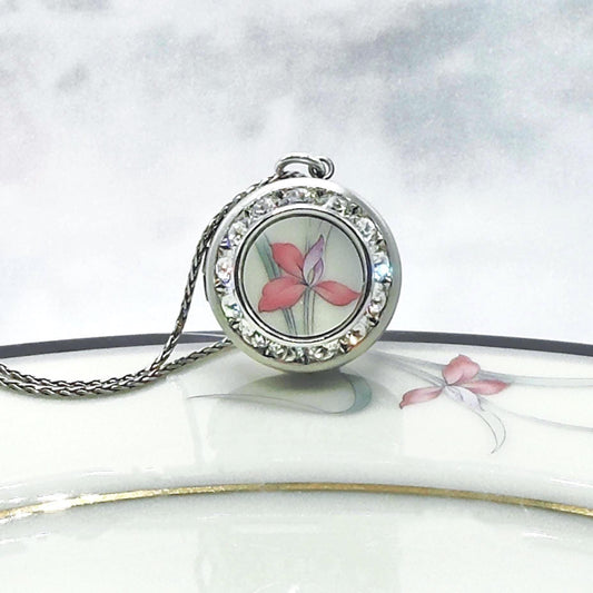 May include: A silver-toned pendant necklace featuring a round locket with a floral design. The locket is adorned with clear crystals around the edge. The floral design inside the locket showcases a pink and purple flower. The necklace chain is also silver-toned.