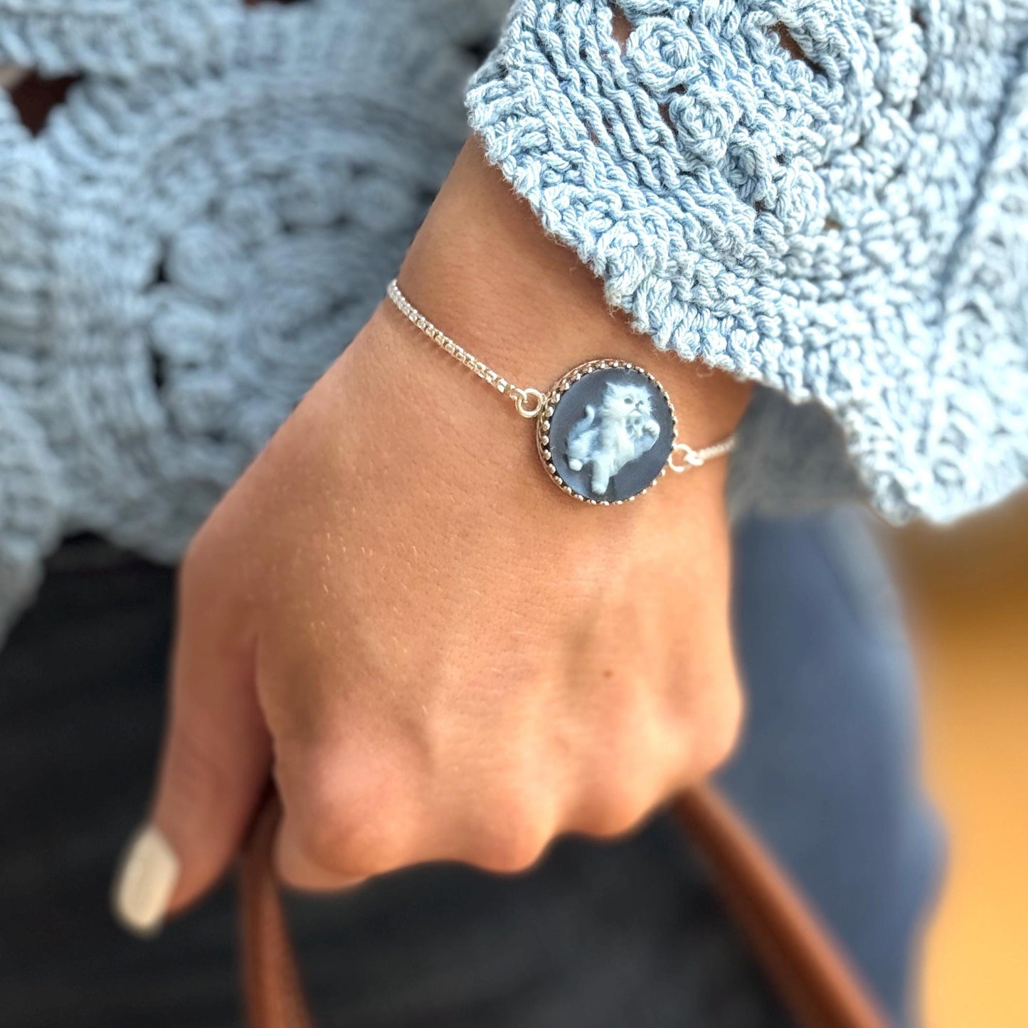 May include: A silver bracelet featuring a round cameo pendant with a light blue cat design. The bracelet is worn on a wrist, with a light blue crocheted sleeve in the background.