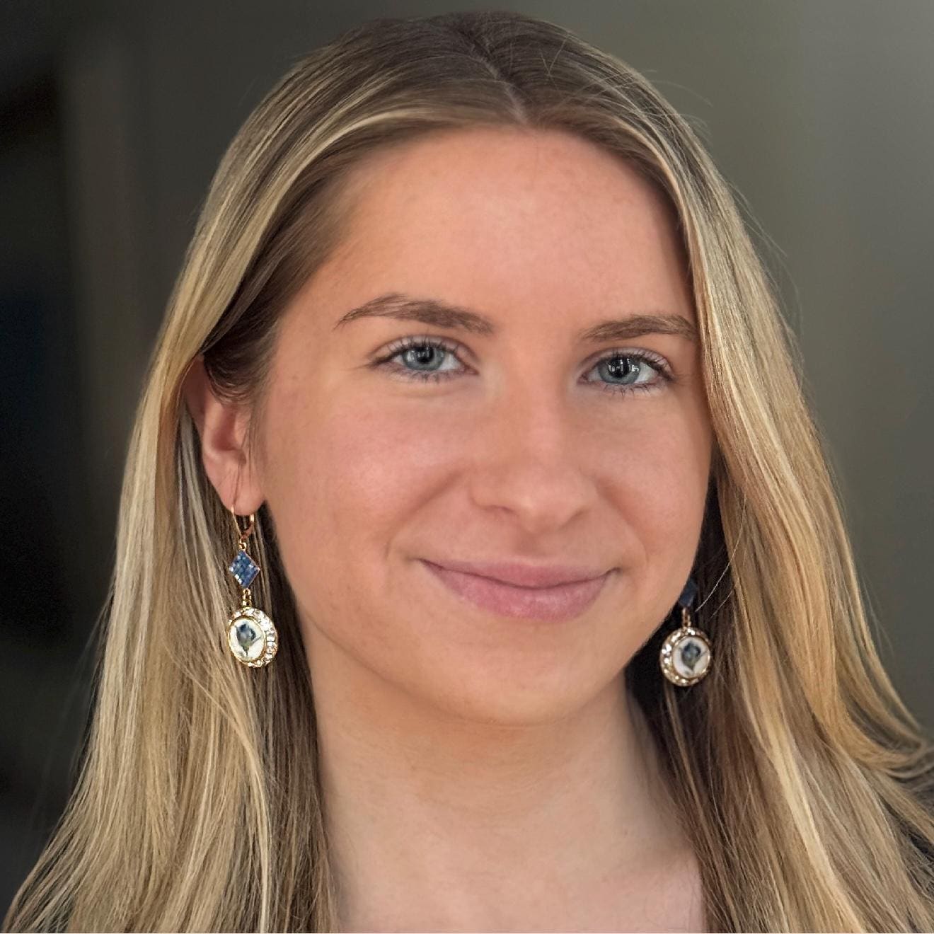 May include: Gold-colored dangle earrings with a blue diamond-shaped stone and a round pendant with a white and blue design. The earrings are worn by a person with blonde hair and blue eyes.