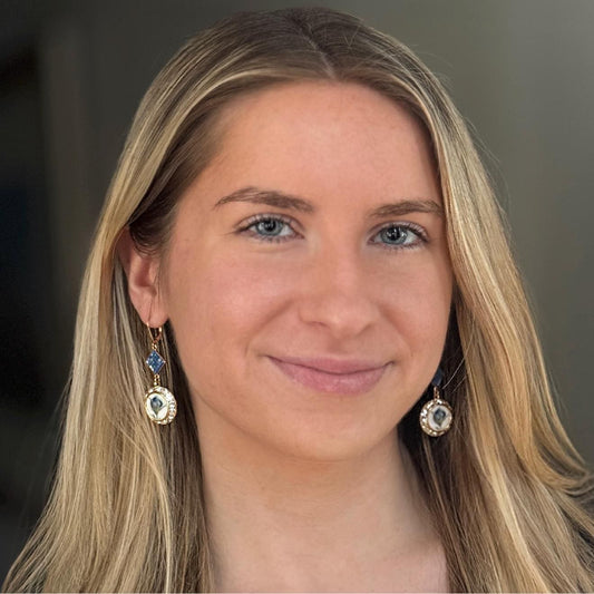 May include: Gold-colored dangle earrings with a blue diamond-shaped stone and a round pendant with a white and blue design. The earrings are worn by a person with blonde hair and blue eyes.