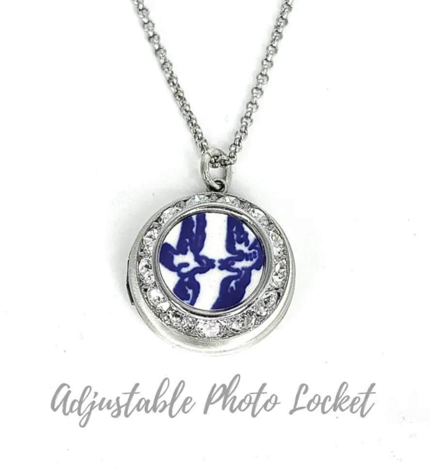 May include: A silver-toned circular locket necklace featuring a central inlay with a blue and white abstract pattern, surrounded by a border of clear crystals. The locket hangs from a delicate silver-toned chain. The text Adjustable Photo Locket is visible.