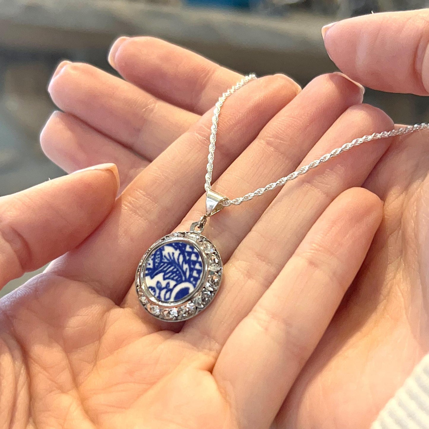 May include: A silver necklace with a round pendant. The pendant features a blue and white floral design surrounded by clear crystals. The necklace is held in open hands.