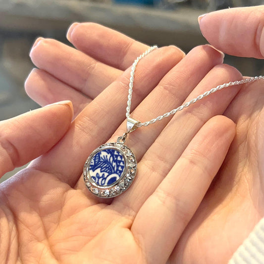 May include: A silver necklace with a round pendant. The pendant features a blue and white floral design surrounded by clear crystals. The necklace is held in open hands.