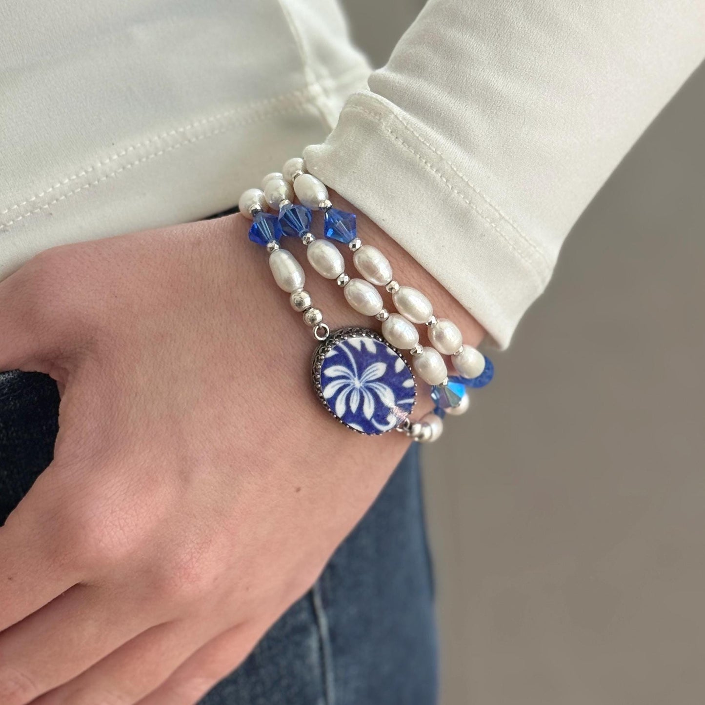 May include: A multi-strand bracelet featuring white pearls and blue crystal beads. The bracelet includes a central oval charm with a blue and white floral design. The jewelry is worn on a wrist, showcasing the elegant design.
