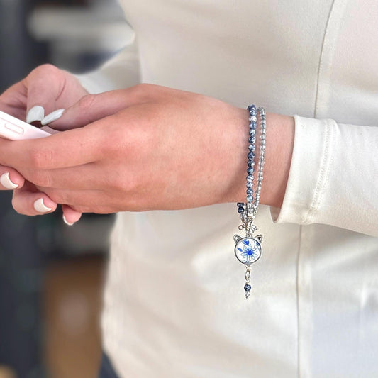 May include: A two-strand bracelet with blue and white beads and a cat-shaped charm. The charm features a blue floral design on a white background. The bracelet is worn on a wrist, with a white long-sleeved shirt in the background.