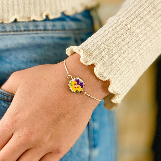 May include: A delicate silver bracelet featuring a round pendant with a pressed pansy flower. The flower has yellow and purple petals. The bracelet is worn on a wrist, with a cream-colored long-sleeved shirt and blue jeans in the background.