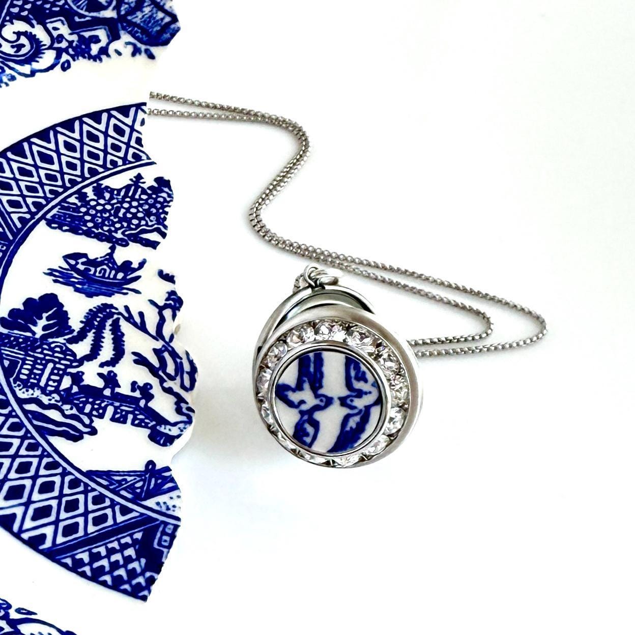 May include: A silver-toned pendant necklace featuring a circular centerpiece with a blue and white bird pattern, surrounded by a ring of clear crystals. The pendant is attached to a delicate silver-toned chain and rests near a blue and white ceramic shard.