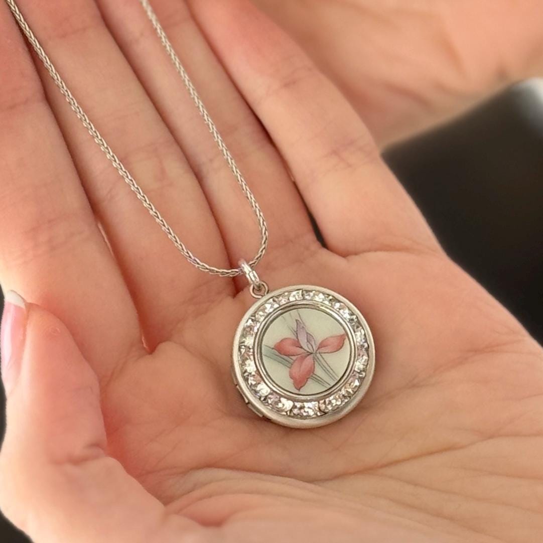 May include: Silver-toned necklace with a round pendant. The pendant features a floral design with a red and purple flower, surrounded by clear crystals. The necklace is held in an open hand.