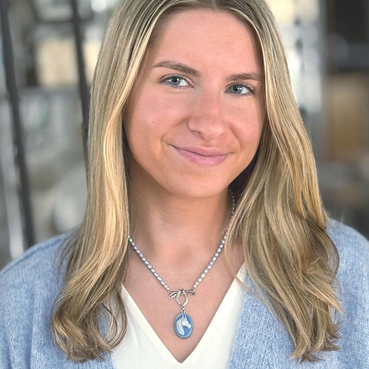 May include: A silver necklace with a blue and white oval pendant featuring a horse head design. The necklace has a pearl-like beaded chain and a bow-shaped clasp. The woman is wearing a light blue sweater.