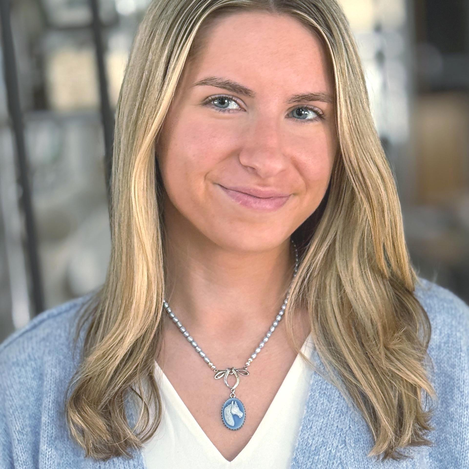 May include: A silver necklace with a blue and white oval pendant featuring a horse head design. The necklace has a pearl-like beaded chain and a bow-shaped clasp. The woman is wearing a light blue sweater.