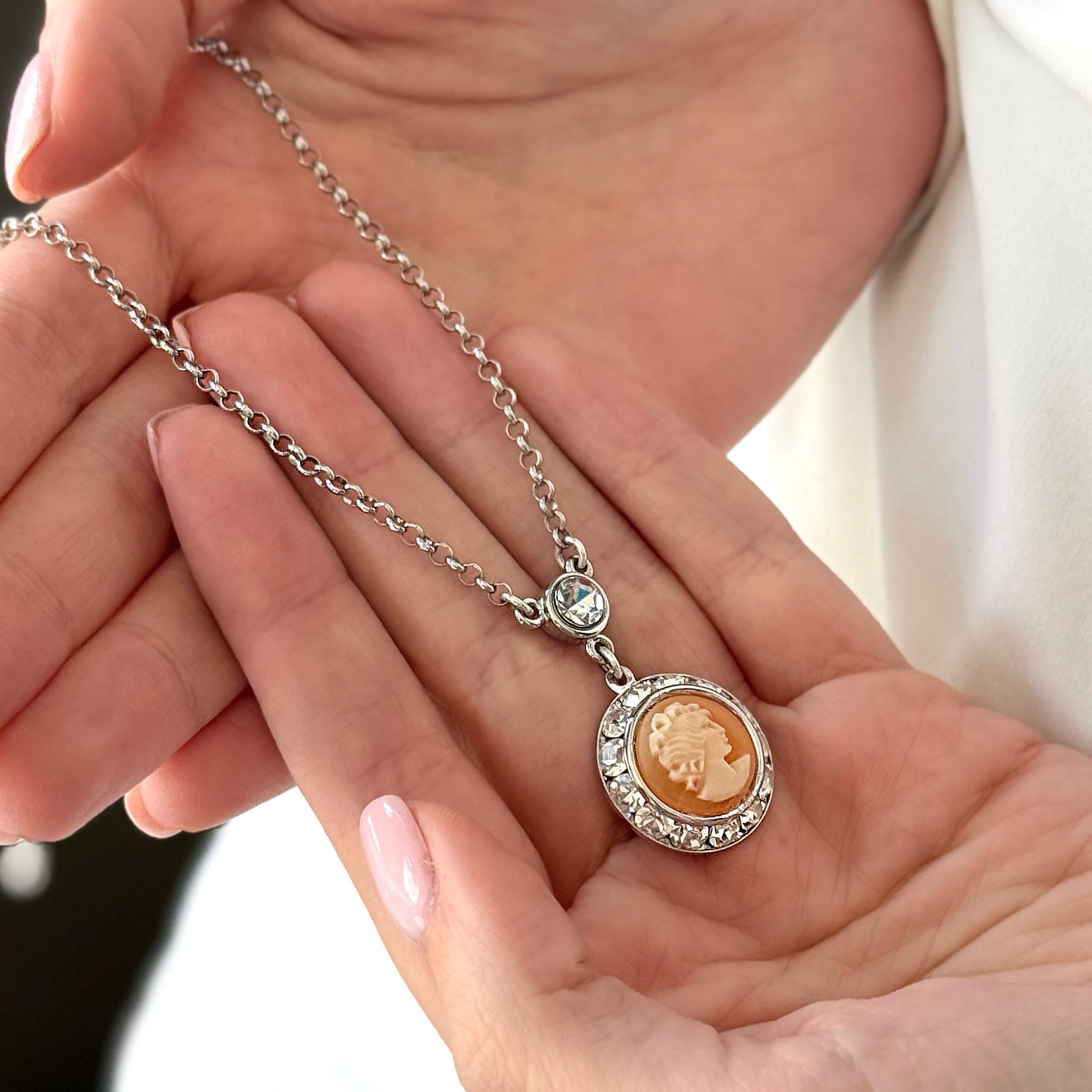 May include: A silver-toned necklace featuring a cameo pendant. The pendant is oval-shaped with a light orange profile and surrounded by clear crystals. A single clear crystal accents the chain above the pendant.