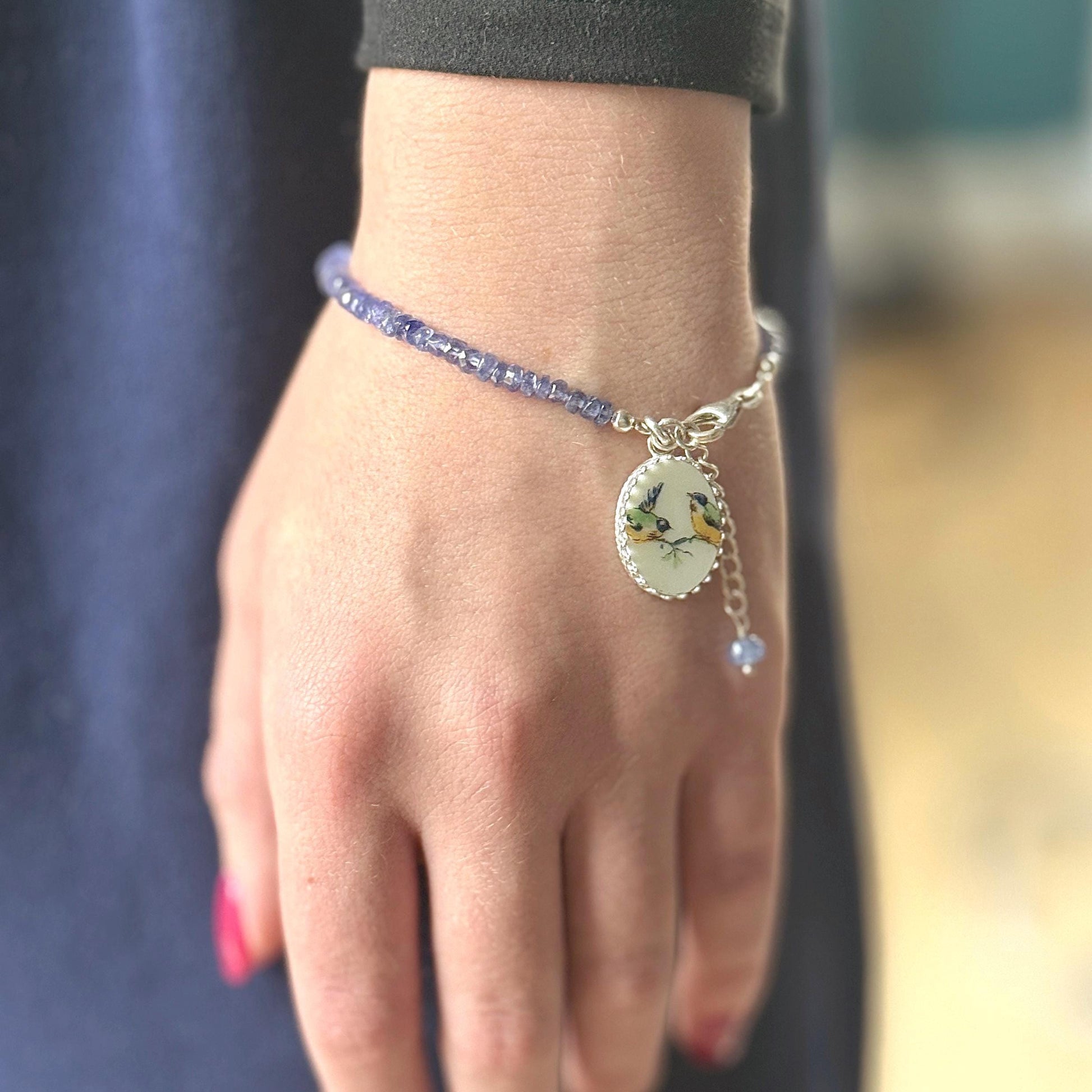 May include: A delicate bracelet featuring small, round, blue beads. A silver charm with a painted oval pendant displaying two yellow and blue birds hangs from the bracelet. A small blue bead dangles below the charm.